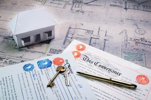 A paper house rests on a blueprint. Next to the house are mortgage papers.