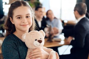 A girl holding a bear with adults talking in the background.